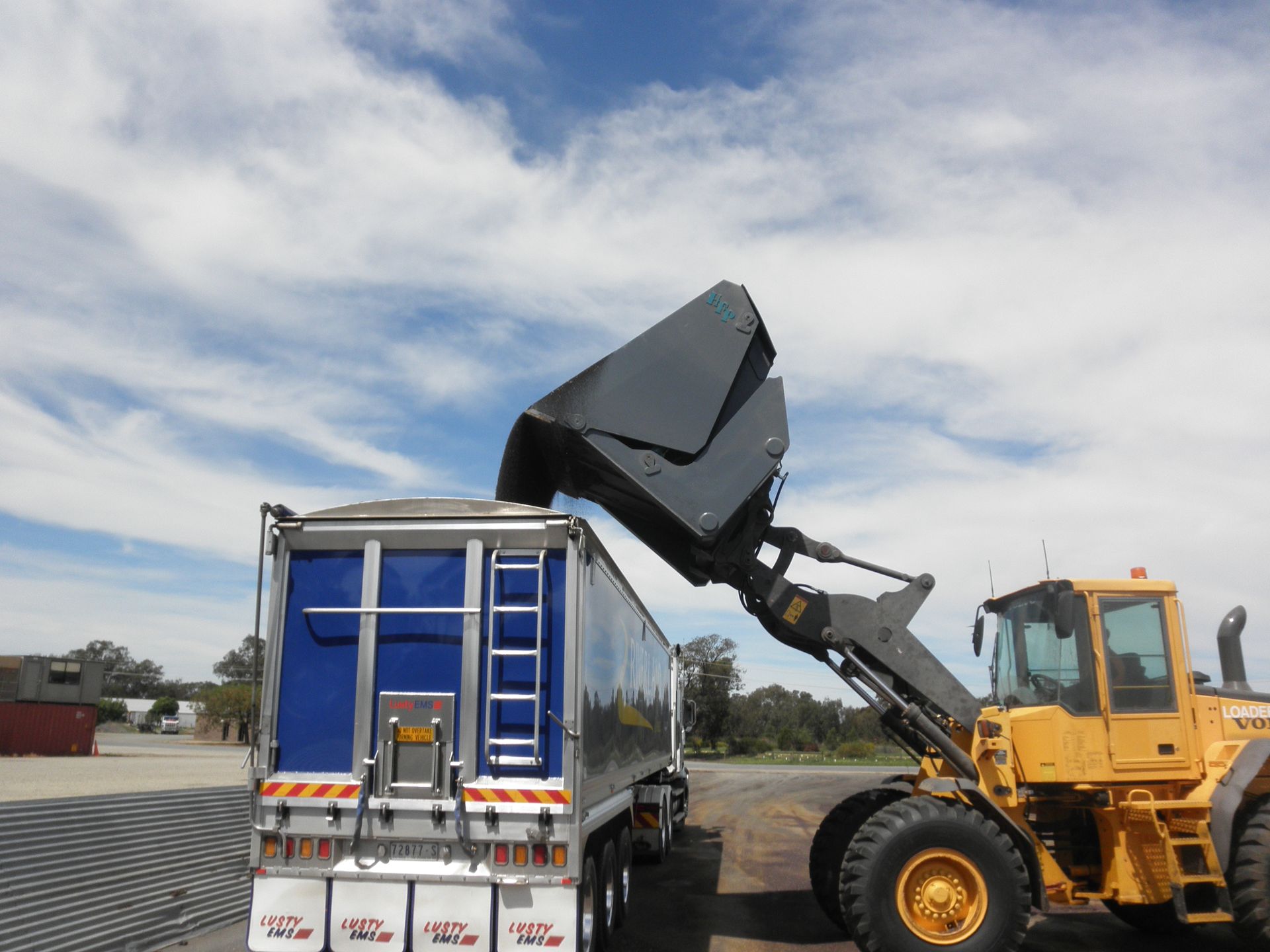 A blue truck is being loaded by a yellow tractor