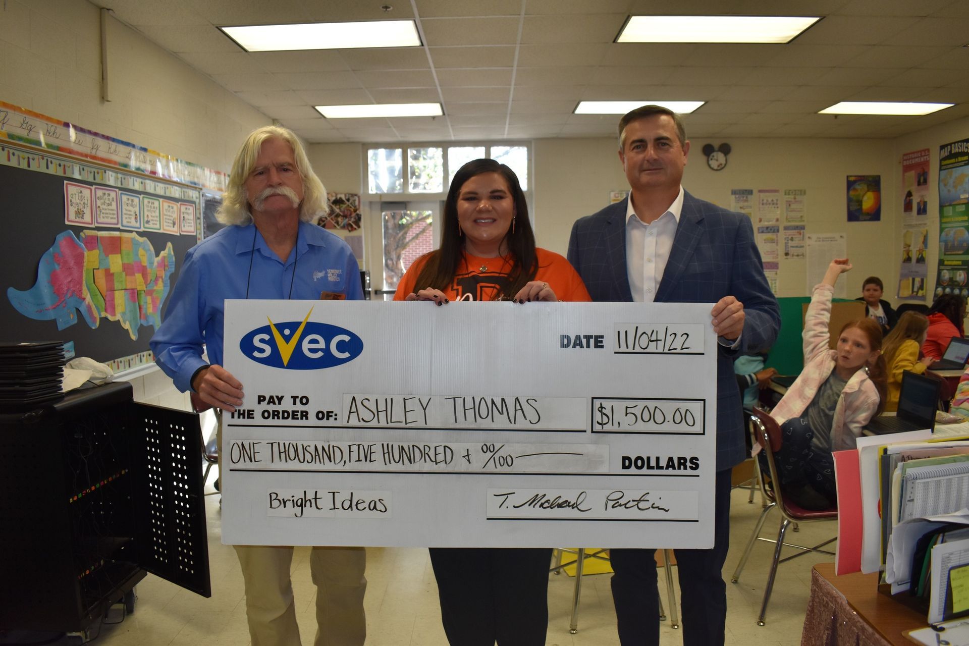 Three people hold a large check in a classroom.