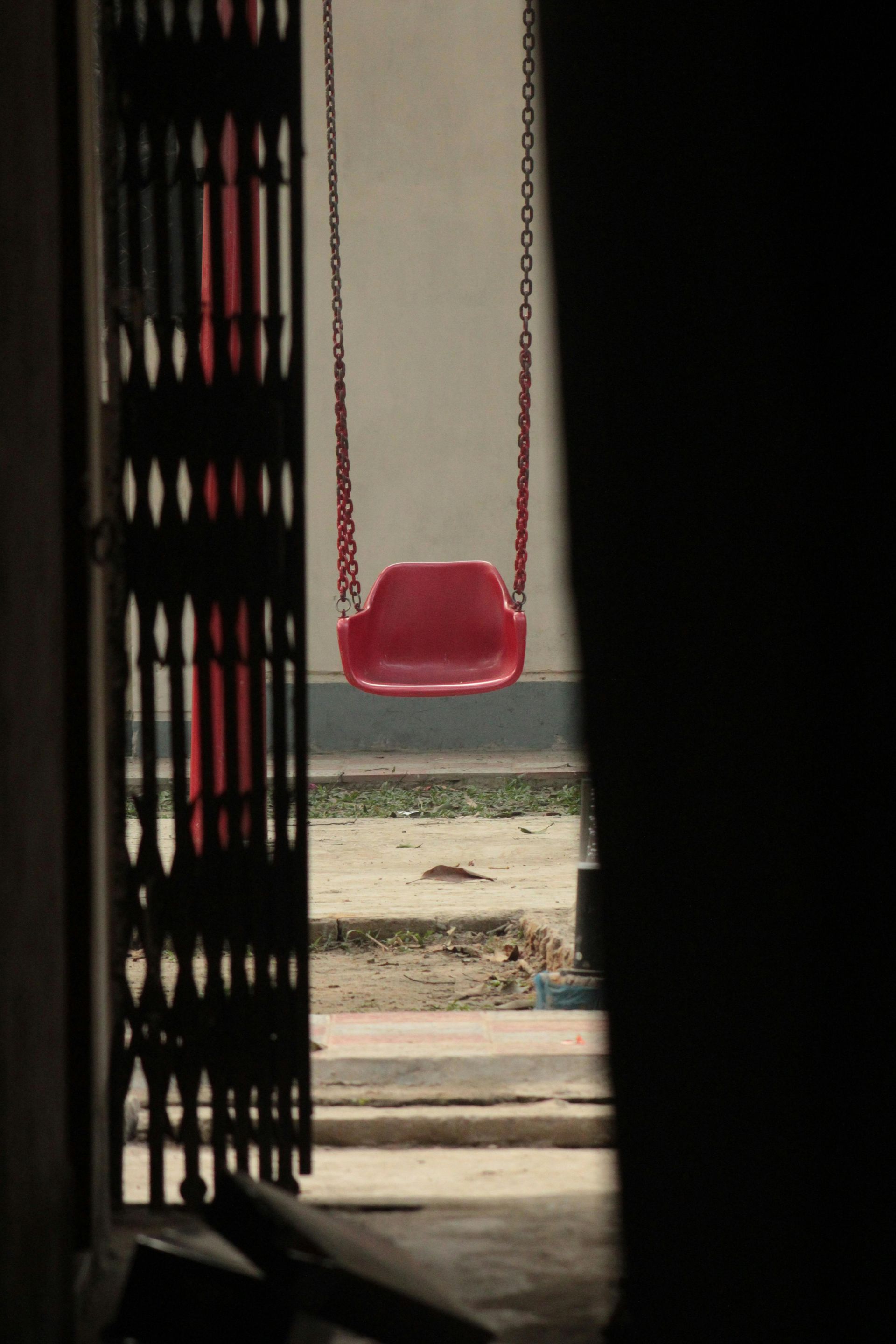 A red swing hangs from chains, viewed through a partly open, dark gate. The background shows a building and ground.