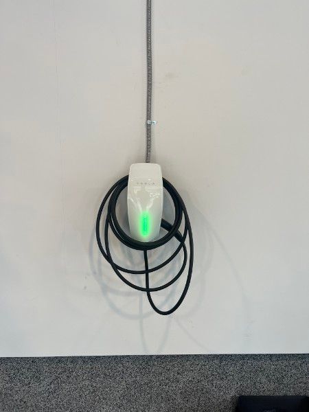 A EV wall charger with a green light on it is hanging from a wire on a wall.