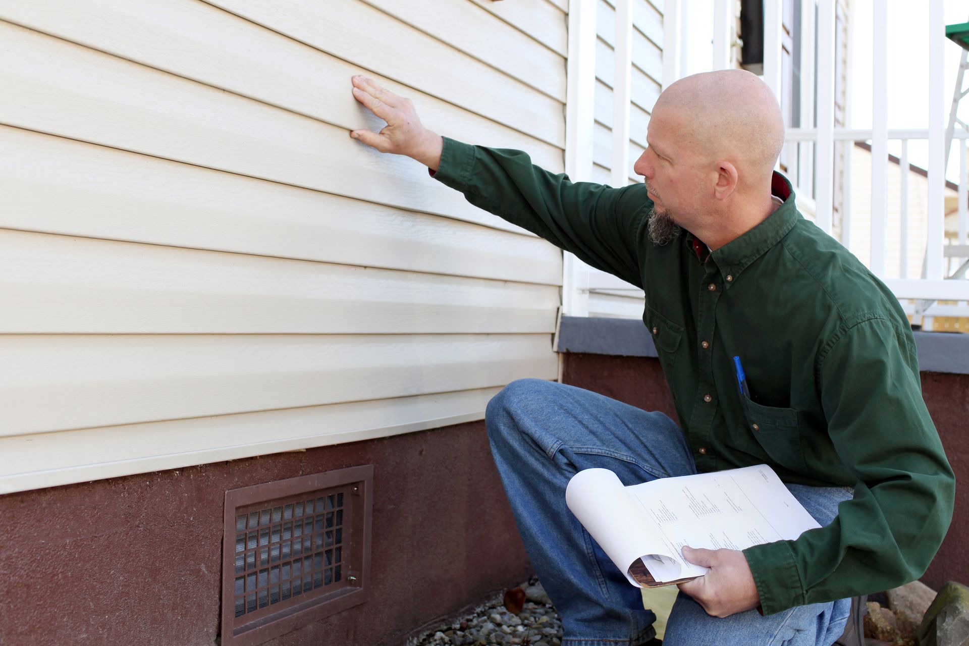 A man is kneeling down in front of a house looking at the siding.