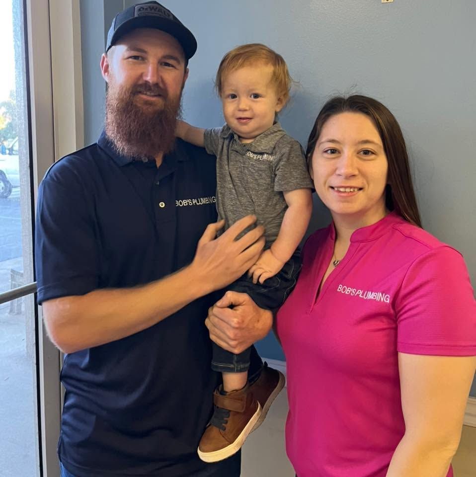 Three people posing indoors, one holding a toddler, wearing branded shirts and smiling