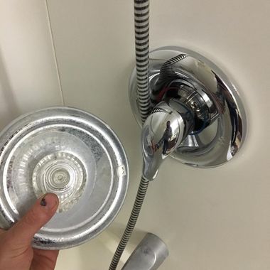 Hand holding a silver shower head attached to a chrome wall mount in a bathroom.
