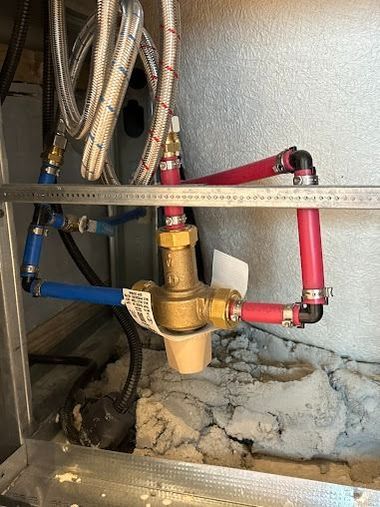 Water heater plumbing with red and blue shutoff valves and flexible braided hoses above a dusty floor.