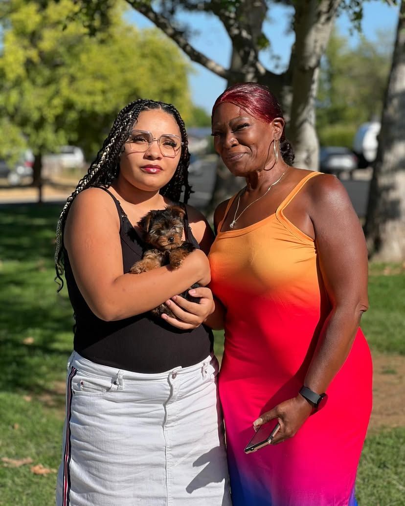 Two women are standing next to each other in a park holding a puppy.