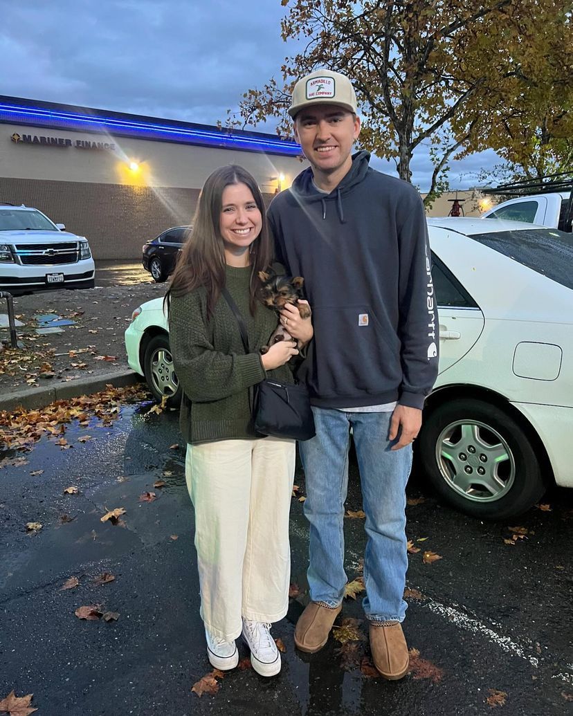 A man and a woman are standing next to each other in a parking lot holding a puppy.