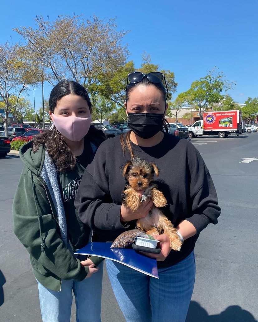 Two women wearing face masks are holding a puppy in a parking lot.