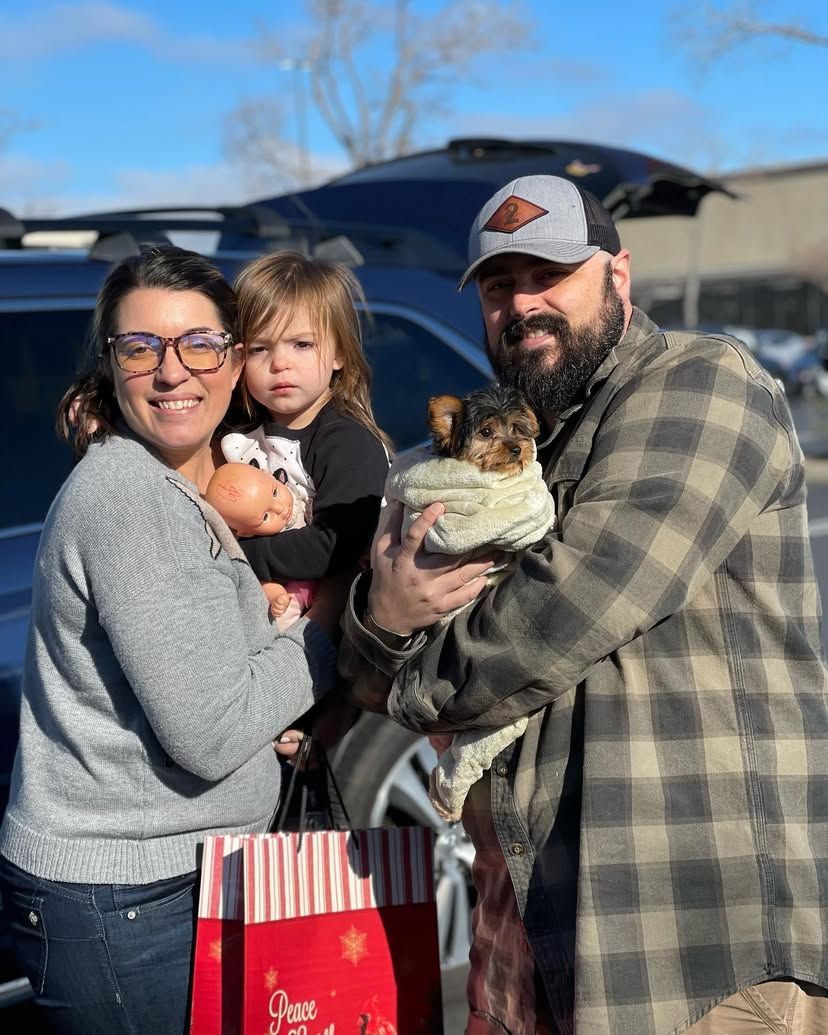 A man and woman are holding a child and a dog in front of a car.