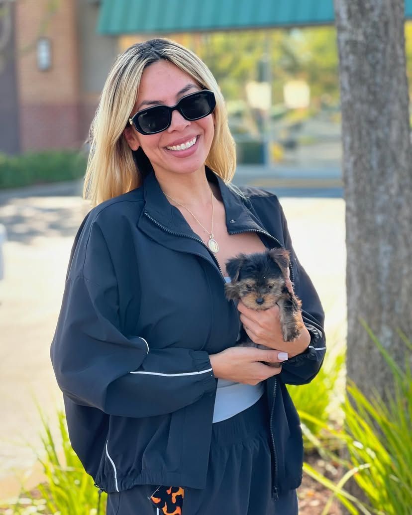 A woman wearing sunglasses is holding a small dog in her arms.