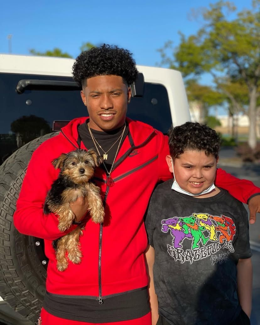 A man in a red jacket is holding a small dog next to a boy in a black shirt.