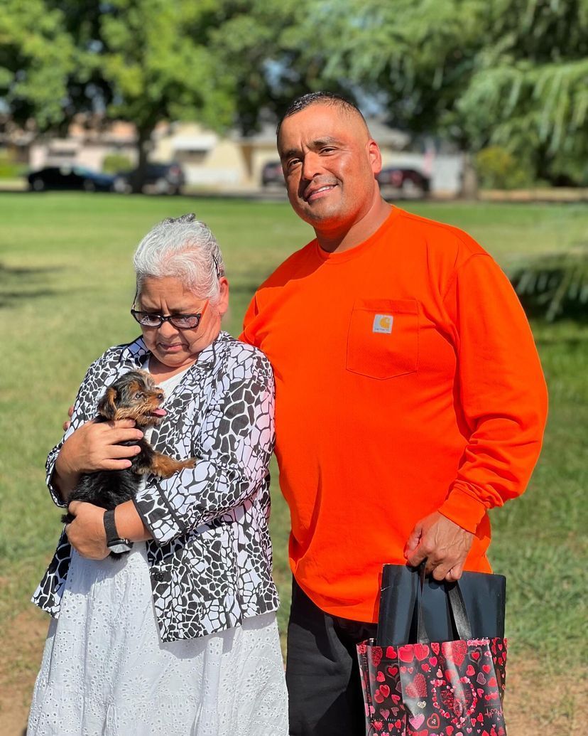 A man in an orange shirt is standing next to a woman holding a small dog.