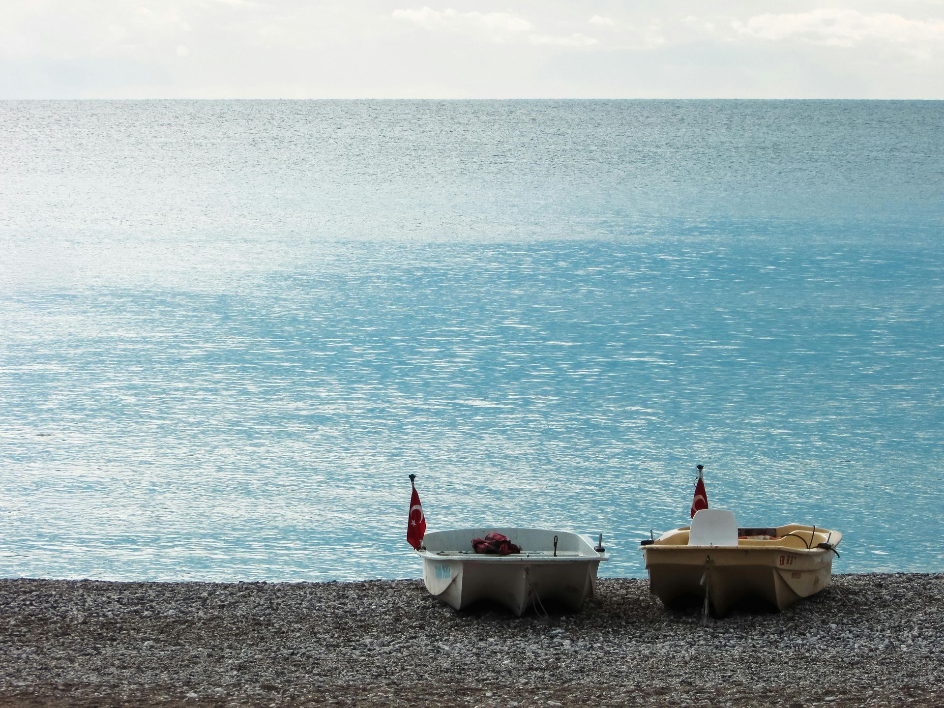 Two small boats on a pebble beach with a sparkling blue ocean under an overcast sky.