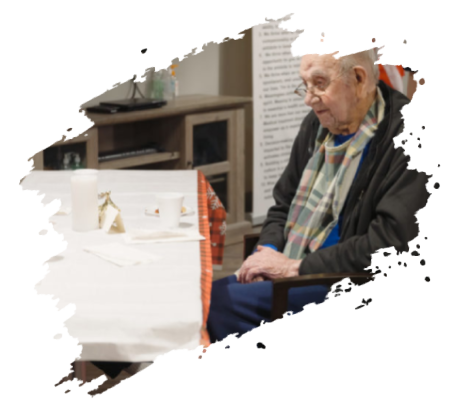 Elderly man in scarf sits at table, looking down. White table, cabinet in background.