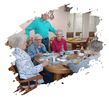 A group of seniors eating at a round table. A man in a turquoise shirt stands nearby.