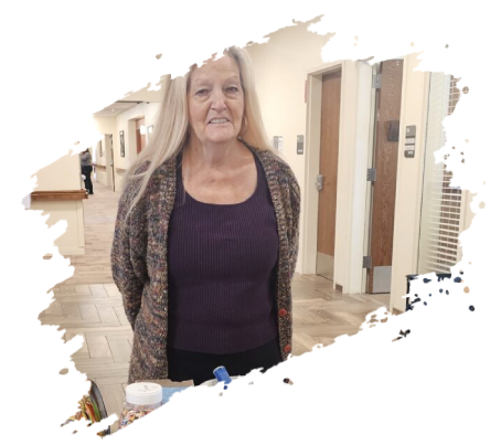 Woman with long blonde hair, purple top, and patterned sweater stands in a hallway near elevators.