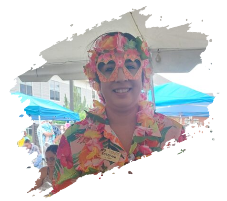 Person in floral shirt, heart-shaped sunglasses, and lei smiles at an outdoor event with tents.