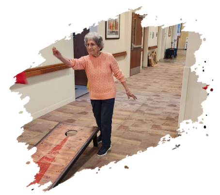 Woman playing cornhole in a hallway, aiming toward the hole, wearing a peach sweater and dark pants.