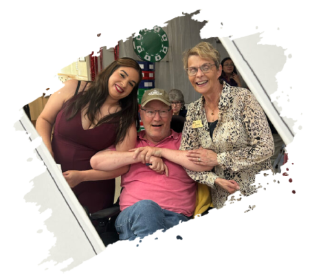 Three people smiling for a photo. A woman in a red dress and a woman in a blazer flank a man in a wheelchair.