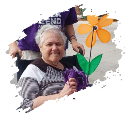 Older woman in wheelchair with flower prop, smiling, someone behind her in purple shirt.