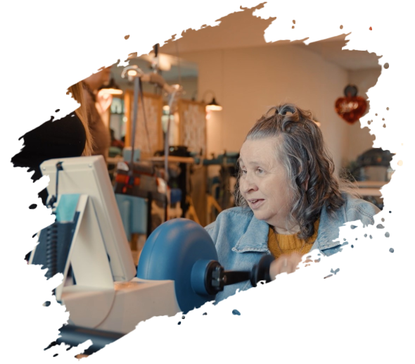 Woman using a blue machine, possibly a lathe, in a workshop setting. She has gray hair and is focused.