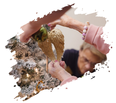 Hands decorating a Christmas tree with golden angel wings. Elderly woman in background.