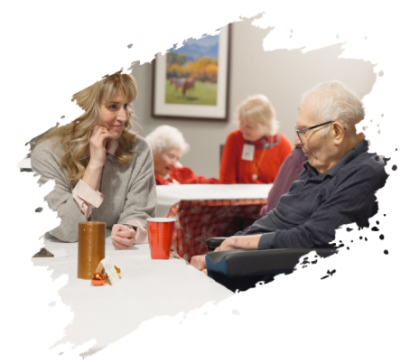 Woman talking to elderly man in a care home setting, others visible.