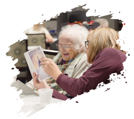 Elderly woman and woman look at a pamphlet, smiling. Indoors. Other people and hats in background.