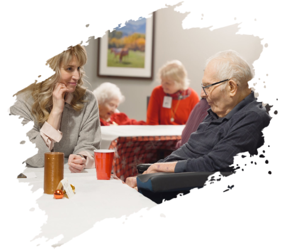 Woman talks to elderly man in a wheelchair at a table; other seniors in background.