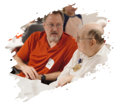 Two men in conversation indoors; one in orange shirt, the other in a white shirt.