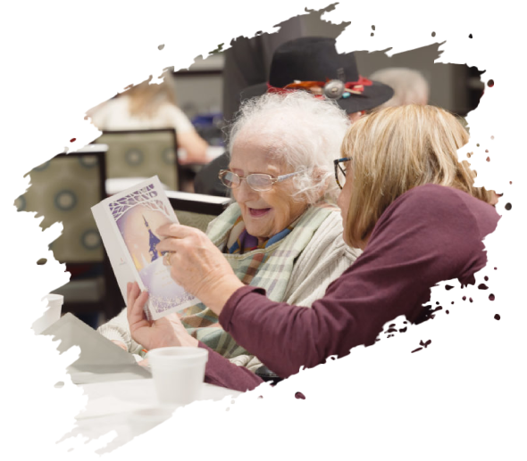 Woman showing a book to an elderly woman. They both smile. Indoors.