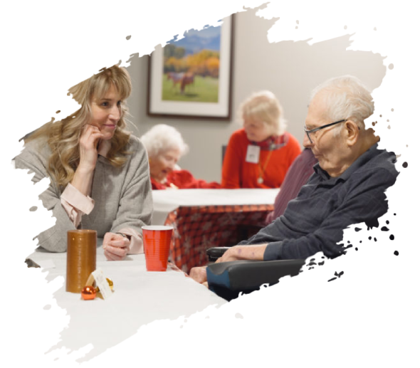 Woman chats with an elderly man in a wheelchair at a table; other people are in the background at a social gathering.