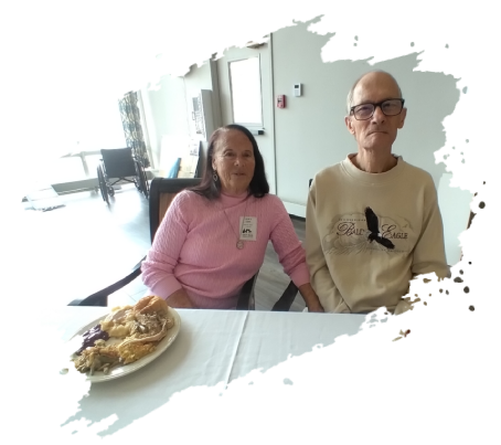 Two seniors sit at a table. Woman in pink smiles. Man in beige sweater, glasses. Plate of food. Bright room.