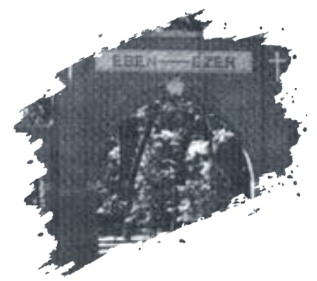 Elderly man wearing glasses and a blue sweater with a white collar, inside a home setting.