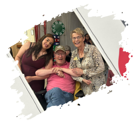 Three people smiling indoors: a man in a wheelchair, two women flanking him. Bright lighting.