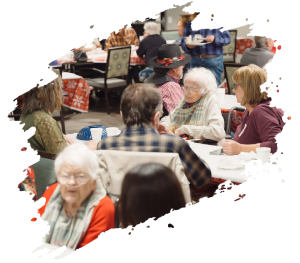 People seated at tables, socializing. Many are older, in a brightly lit room. Some have food, others are talking.