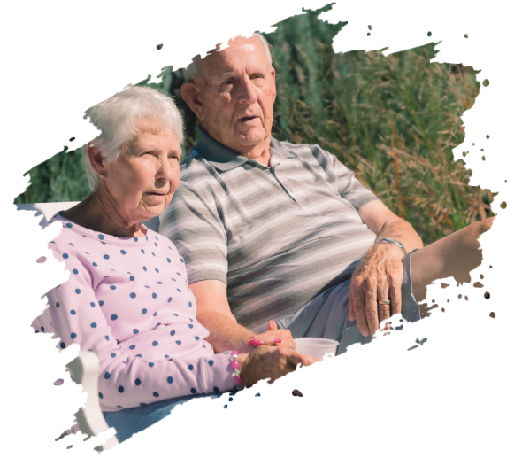 Elderly couple sits outdoors, holding hands. Woman in pink, man in grey, sunny setting.