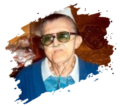 Elderly man wearing glasses and a blue sweater with a white collar, inside a home setting.