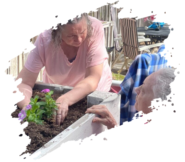 Woman planting flowers with another person nearby in a sunny garden setting.
