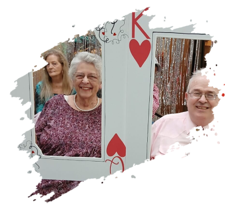 Three people smiling, posing behind a playing card cutout, decorated with hearts.