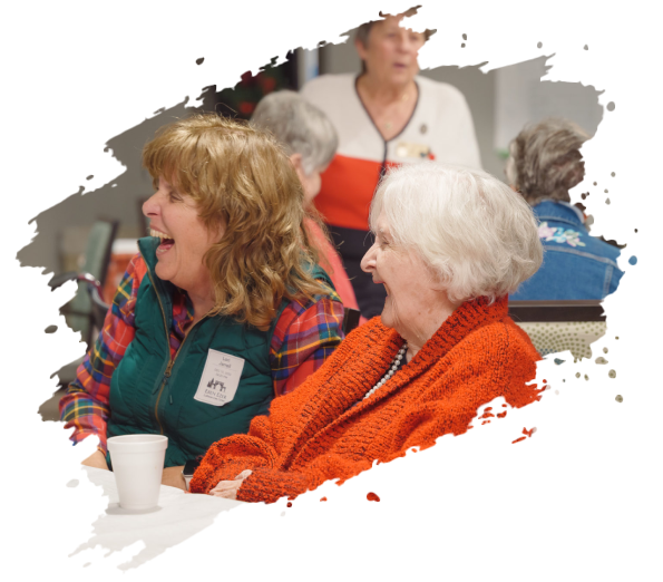 Two women laughing in a room; one in orange, one in plaid with a green vest. Others are nearby.