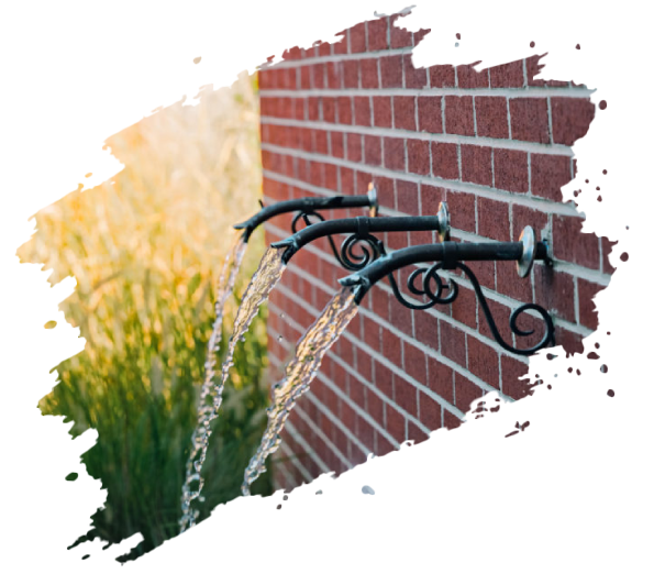Water fountain with three spouts against a brick wall, water flowing, gold-colored grass in the background.