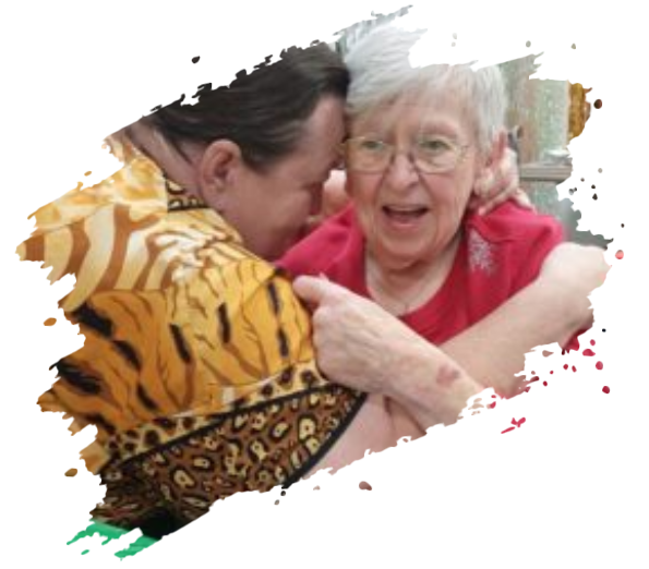 Woman in red shirt hugs elderly woman, both smiling. Tiger-print shirt visible, indoor setting.