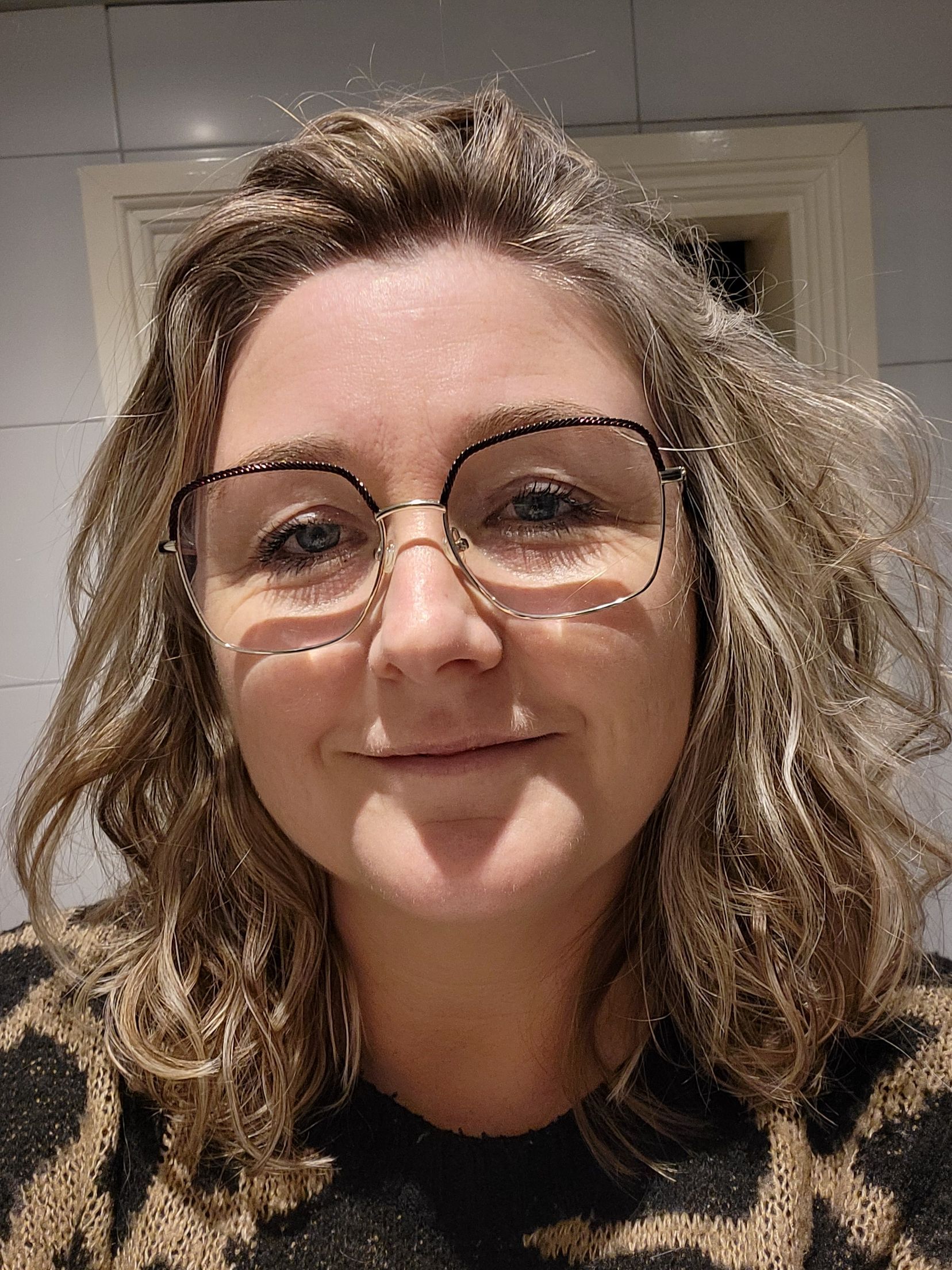 Een vrouw met krullend haar, een bril en een top met print kijkt lachend naar de camera en staat afgebeeld tegen een lichtgekleurde betegelde muur.