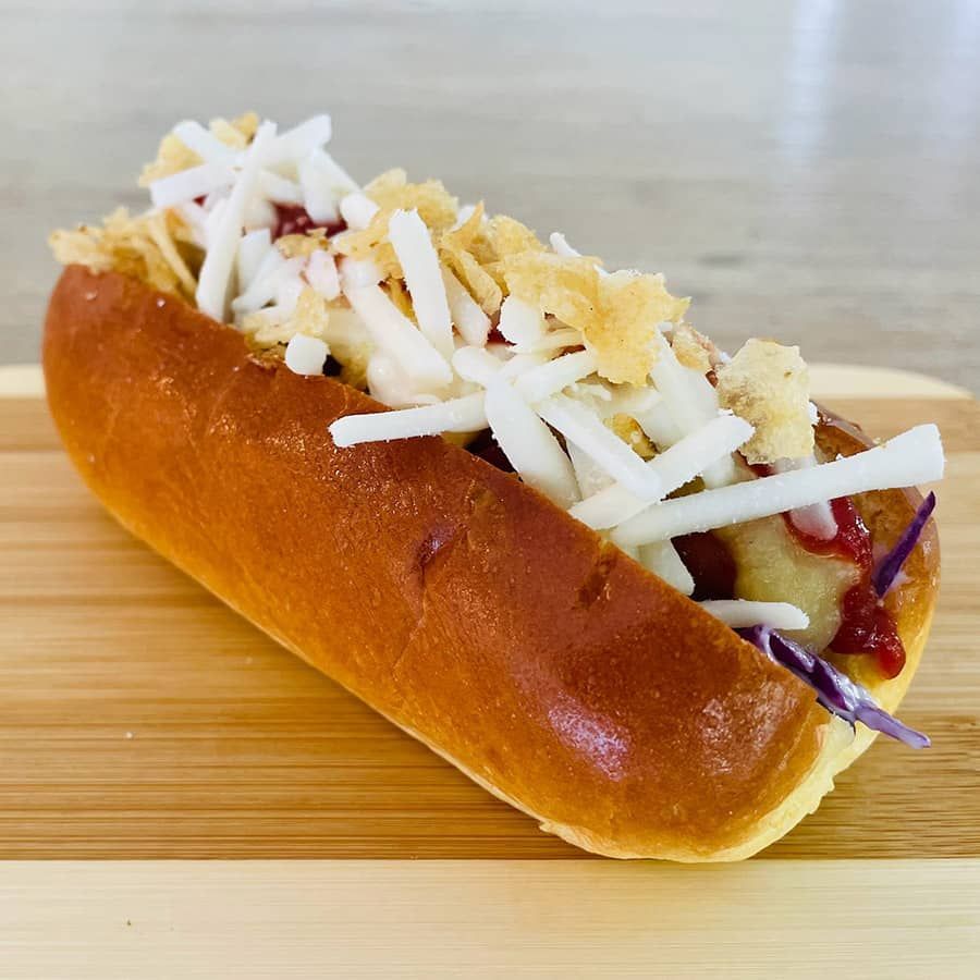 A hot dog with coleslaw and cheese on a bun on a wooden cutting board.