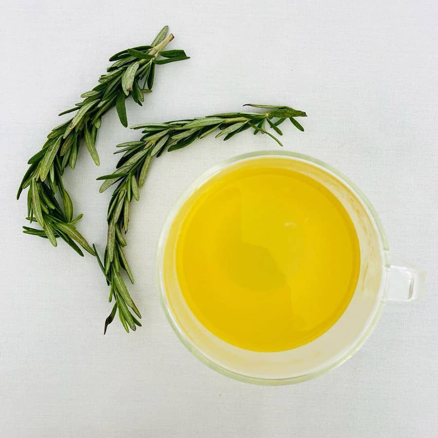 A cup of oil next to a sprig of rosemary