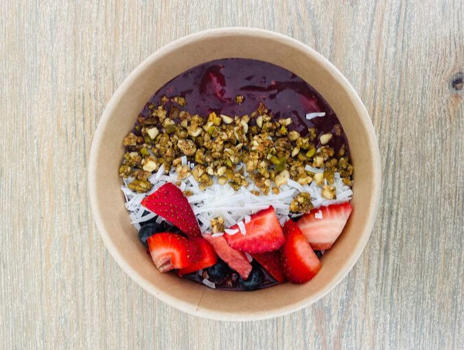 A bowl of acai bowl with strawberries , blueberries , granola and coconut on a wooden table.