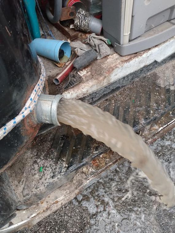 Dirty water pouring from a pipe onto a wet concrete floor near debris and equipment.