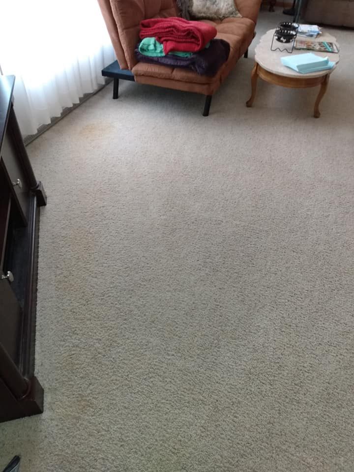 Small living room corner with beige carpet, chairs, a round table, and folded clothes by a window.