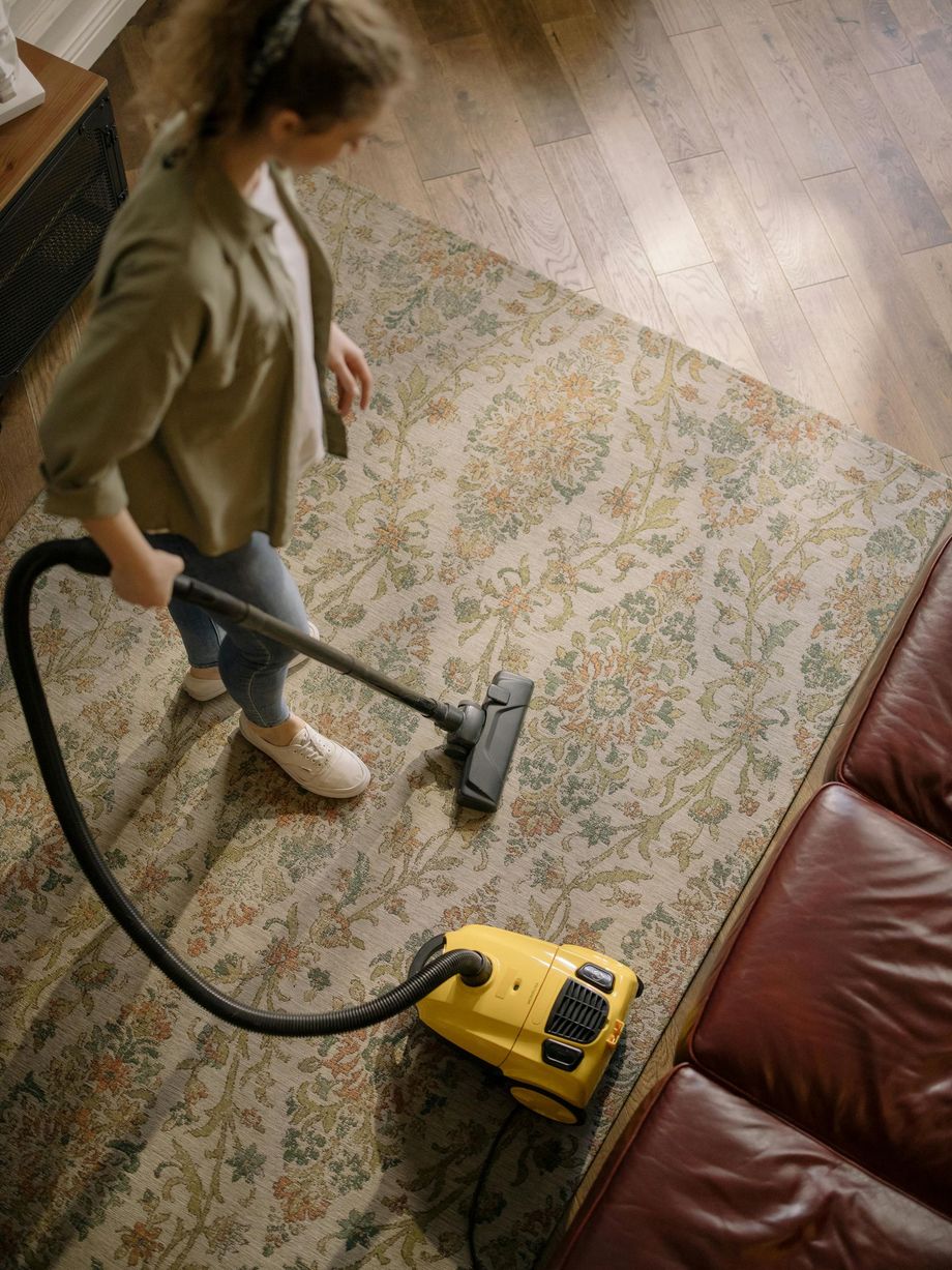 Person vacuuming a patterned rug beside a brown couch with a yellow vacuum cleaner.