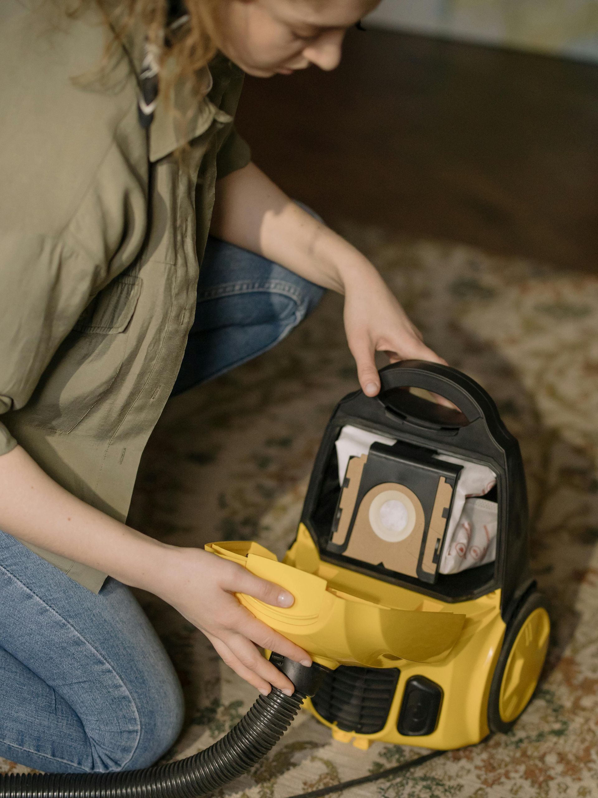 Person kneels beside a yellow canister vacuum, adjusting its dust compartment on a carpeted floor
